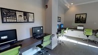 This single-floor business center is designed to provide a functional and comfortable workspace. The room features a series of wall-mounted, dark wood-finished desks that run along the perimeter and extend around a central structural column, creating distinct yet open workstation areas. Each station is equipped with a desktop computer and keyboard, offering residents or guests convenient access to technology.

The seating consists of modern green ergonomic chairs with chrome bases, adding a pop of color against the neutral gray walls and light carpeted flooring. Recessed lighting and natural light filtering in from one side of the room keep the space bright and welcoming.

Decorative framed artwork with a vintage typographic theme adorns the walls, adding visual interest without overwhelming the professional atmosphere. The layout is thoughtfully organized to maximize available space while maintaining an open, uncluttered feel. The overall ambiance is clean, contemporary, and practical — well-suited for tasks such as remote work, printing, or browsing, making it a convenient amenity within the larger property.