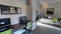 This single-floor business center is designed to provide a functional and comfortable workspace. The room features a series of wall-mounted, dark wood-finished desks that run along the perimeter and extend around a central structural column, creating distinct yet open workstation areas. Each station is equipped with a desktop computer and keyboard, offering residents or guests convenient access to technology.

The seating consists of modern green ergonomic chairs with chrome bases, adding a pop of color against the neutral gray walls and light carpeted flooring. Recessed lighting and natural light filtering in from one side of the room keep the space bright and welcoming.

Decorative framed artwork with a vintage typographic theme adorns the walls, adding visual interest without overwhelming the professional atmosphere. The layout is thoughtfully organized to maximize available space while maintaining an open, uncluttered feel. The overall ambiance is clean, contemporary, and practical — well-suited for tasks such as remote work, printing, or browsing, making it a convenient amenity within the larger property.