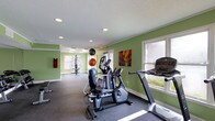This dedicated fitness center offers a comprehensive workout environment designed to support a variety of exercise routines. The space is finished with bold sage green walls that create an energizing atmosphere, complemented by a neutral gray rubber flooring ideal for high-traffic athletic use. Recessed lighting throughout ensures consistent, even illumination, while multiple windows along the walls invite ample natural light and views of the surrounding greenery, contributing to an open and airy feel. The fitness floor is outfitted with a range of cardio equipment, including a treadmill, elliptical trainer, and a recumbent stationary bike, providing options for low- and high-impact aerobic workouts. Toward the rear of the space, a multi-function cable and weight machine offers opportunities for strength training and resistance exercises. Exercise balls are also on hand for core and flexibility training. Colorful wall art adds a decorative touch, keeping the environment visually engaging. A textured white ceiling with recessed can lights ties the room together with a clean, finished look. The layout allows for comfortable movement between stations, making this fitness center a functional and well-organized space suitable for a broad range of fitness activities.