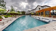 Located at 4700 Fossil Dr in Haltom City, Texas, this outdoor amenity space is a thoughtfully designed resort-style retreat that serves as a central gathering hub for its community. The centerpiece is a generously sized, freeform swimming pool with crystal-clear blue water, featuring built-in sun shelves with in-water loungers and gentle water features that add both visual appeal and a relaxing ambiance. The pool deck is finished with a sophisticated patterned paver surface that extends throughout the outdoor area, providing a clean and durable foundation for all activities. A striking warm-toned wood pergola anchors one corner of the pool deck, offering a shaded social space ideal for outdoor dining or casual gatherings. Adjacent to the pool, lush green lawn areas provide additional lounging space, lined with modern white chaise lounges, vibrant green umbrellas, and colorful accent furniture. Mature trees and native landscaping add privacy and natural beauty, while raised planting beds with ornamental grasses and shrubs contribute to the polished, layered aesthetic. The surrounding multi-story residential buildings feature a contemporary design combining white board-and-batten siding with brick accents, framing the outdoor space with an architecturally cohesive backdrop. The overall atmosphere is upscale, inviting, and well-appointed for outdoor relaxation and socializing.