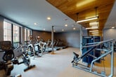 This modern fitness center is thoughtfully designed to support a full range of workout routines in a spacious, open-plan layout. The interior is defined by a striking combination of warm wood ceiling panels and cool slate-blue walls, creating a contemporary aesthetic that balances energy and focus. Recessed lighting throughout, supplemented by sleek pendant fixtures, ensures the space is well-lit at all times.

The cardio zone is generously outfitted with a row of treadmills, elliptical trainers, and upright cycling bikes, all positioned along large windows that allow natural light to filter in and offer views of the surrounding exterior. A dedicated strength training area features a Matrix rowing machine, a functional cable cross trainer, adjustable weight benches, and a full dumbbell rack organized for easy access. A wall-mounted flat-screen television is positioned above the free weights area for added convenience.

Full-length mirrors line the strength training walls, visually expanding the space and allowing users to monitor their form during exercises. A glass-paneled partition separates sections of the facility, contributing to the open yet organized feel. Amenities include a built-in water fountain, ensuring hydration is always within reach. The durable carpet flooring throughout provides comfort and traction, making this fitness center a practical and polished facility.