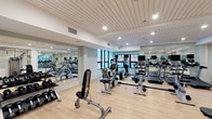 This fitness center is thoughtfully designed to accommodate a wide range of exercise routines within a bright, modern environment. The main workout floor features light hardwood-style flooring, a slatted white ceiling with recessed lighting, and large windows that allow natural light to fill the space while offering views of the surrounding urban streetscape. Floor-to-ceiling mirrors line the walls, visually expanding the room and allowing users to monitor their form. The area is outfitted with an extensive array of cardio equipment, including multiple treadmills and stationary bikes with integrated display screens, alongside strength training machines, free weights, adjustable benches, and cable systems. A dedicated studio room, enclosed by black-framed glass partitions, provides a separate zone for spinning bikes, balance balls, and functional training accessories. A built-in storage station with open wood shelving, white cabinetry, and a quartz countertop offers convenient organization near the gym floor. A wall-mounted hydration station with a bottle-filling feature is positioned near the entrance for easy access. Together, these elements create a clean, functional, and professional fitness environment.