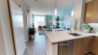 This single-floor unit showcases a thoughtfully designed open-concept layout that seamlessly integrates the kitchen and living areas. The kitchen features a generous white quartz island with an undermount sink, warm wood cabinetry, stainless steel appliances including a built-in dishwasher, and elegant pendant lighting suspended overhead. A sliding barn door near the entry adds a contemporary architectural detail while maximizing usable floor space.

The living area is defined by striking teal accent walls that create a bold, energetic contrast against the crisp white ceilings and neutral-toned luxury vinyl plank flooring that runs throughout. A large window floods the space with natural light and provides outward views, enhancing the open, airy feel of the room. The living space comfortably accommodates a full seating arrangement, with room for accent chairs, a sofa, and a central coffee table atop a textured area rug. Lush indoor plants are incorporated throughout, adding warmth and a natural element to the modern interior. A wall-mounted split AC unit ensures year-round climate comfort. Overall, the space balances vibrant color with clean, modern finishes.