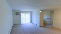 This single-floor unit offers a clean, functional layout centered around a spacious living area. The room is finished with wall-to-wall neutral carpeting that extends throughout, contributing to a quiet and comfortable atmosphere. A large sliding glass door serves as the primary source of natural light, fitted with vertical blinds that allow for adjustable privacy and light control. The door provides a direct view of outdoor greenery, bringing a subtle connection to the outside.

A wall-mounted air conditioning unit is positioned to the left of the sliding door, offering climate control directly within the living space. A baseboard heating unit runs along the lower wall near the door, providing an additional heating option. The walls and ceiling are finished in a clean, neutral white, keeping the space feeling open and bright.

A partial wall or column on the right side of the living area creates a natural visual division, with a built-in shelf visible beyond it, suggesting an adjacent area that could serve as a dining space or entryway. A ceiling-mounted light fixture provides additional illumination in this secondary zone. Overall, the space offers a practical, well-lit environment suited for everyday living.