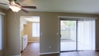 This single-floor residence offers a practical and comfortable layout designed to maximize functionality. The living area features warm hardwood-style flooring and a ceiling fan, creating a relaxed and well-ventilated atmosphere. Neutral, earthy-toned walls provide a versatile backdrop throughout the space.

A sliding glass door in the living area opens to an outdoor patio or balcony, allowing natural light to filter in while providing easy access to the exterior. Vertical blinds offer privacy and light control as needed.

Adjacent to the living area, the kitchen is accessible through an open passageway, promoting a connected and sociable floor plan. The kitchen features a drop ceiling with recessed lighting panels, white cabinetry, and a white range, giving it a clean and classic appearance. A window in the kitchen further enhances the natural light within the space.

The textured ceilings are consistent throughout, adding a subtle architectural detail to the interior. Overall, this single-floor residence combines an open, connected layout with ample natural light, neutral finishes, and practical features, making it a functional and inviting living environment.
