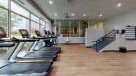 Located at 50 Park Row W in Providence, Rhode Island, this well-appointed fitness and aquatic facility offers a comprehensive wellness experience across a single, thoughtfully organized floor. The gym area is divided into distinct functional zones, each finished with light hardwood-style flooring and recessed lighting throughout. The cardio zone features a row of Matrix treadmills and ellipticals positioned alongside large windows that invite natural light and offer views of the surrounding cityscape. Adjacent areas house a robust selection of strength training equipment, including cable machines, a full dumbbell rack, a weight bench, and a functional training rig with accessories such as a rowing machine, stability balls, and resistance tools. An upright exercise bike occupies a central open area near the main entrance, which is accessed through elegant wood-framed glass doors. The aquatic area showcases a generously sized indoor lap pool with tile surrounds and stone-look flooring, separated from the fitness floor by floor-to-ceiling glass partitions. A relaxing hot tub is set against a striking sculptural wave-textured accent wall, adding a design focal point to the space. Recessed lighting throughout maintains a clean, modern ambiance across all areas of this fully equipped facility.