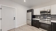 This thoughtfully designed single-floor unit showcases a contemporary aesthetic throughout, blending clean lines with high-quality finishes. The kitchen is a standout feature, equipped with stainless steel appliances including a French door refrigerator, electric range, and over-the-range microwave. A striking combination of dark lower cabinetry and crisp white upper cabinets is complemented by a bold black marble-patterned backsplash and white countertops. A peninsula with a dark base provides additional prep and dining space, accented by a pendant light above. Light-toned luxury vinyl plank flooring runs consistently throughout the unit, enhancing the bright and airy feel of each room. The bedroom is a calm, neutral space bathed in natural light from a window overlooking greenery, with warm beige walls and clean white trim. The bathroom is finished with dark marble-look floor tiles, a marble-surround soaking tub and shower combination, a wall-mounted vanity with a light wood finish, and a frameless mirror. Smart home features are evident, including a keypad entry door lock and a wall-mounted thermostat. The overall design presents a cohesive, modern living environment with attention to detail in every room.