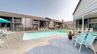 Located at 4020 S 130th East Ave in Tulsa, Oklahoma, this outdoor amenity area centers around a generously sized swimming pool with clear, blue-green water and clearly marked depth indicators along the pool deck. The surrounding concrete deck provides substantial space for relaxation and recreation, lined with an assortment of lounge chairs, tables, and seating arrangements — including aqua-toned chairs and umbrellas — designed to accommodate a large number of guests comfortably.

A standout architectural feature of this space is the gabled pavilion, constructed with a striking exposed wood truss design and a ceiling fan for added comfort during warm months. The pavilion shelters a raised stone-clad structure, which serves as a focal point for the outdoor gathering area. Adjacent to the pavilion, large glass sliding doors connect the pool area to an interior clubhouse or amenity building, allowing for seamless indoor-outdoor flow.

The pool area is enclosed by a black iron fence, providing security while maintaining an open, airy feel. Surrounding residential buildings with multi-level balconies are visible beyond the perimeter, contextualizing the space within a larger community setting. The layout and amenities reflect a thoughtfully designed communal outdoor retreat suited for both leisure and socializing.