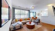 Located at 50 Lily Green Ct NW in Concord, NC, this single-floor residence offers a thoughtfully designed layout filled with natural light and bold, vibrant décor. The open-concept living and dining area is anchored by expansive windows dressed with patterned blue curtains, allowing generous natural light and views of the surrounding greenery. Crown molding and a ceiling fan add classic finishing touches to the living space, which flows seamlessly into a dedicated dining nook featuring a glass-top table, tufted orange chairs, and rustic floating wall shelves. The kitchen is outfitted with white cabinetry, ample counter space, and hardwood-style flooring that carries throughout the main areas. The primary bedroom features a striking teal accent wall, plush carpeting, and a walk-in closet, while the second bedroom offers a cheerful yellow-green accent wall and bright natural light. Both bathrooms include white vanity cabinetry, large mirrors with Hollywood-style lighting, and hardwood-style flooring. The overall ambiance is energetic and welcoming, with cohesive use of color and contemporary furnishings that give the space a lively, modern character.
