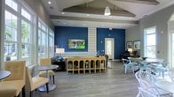 This well-appointed single-floor space exudes a modern, inviting atmosphere designed to encourage gathering and relaxation. The room is anchored by striking cobalt blue accent walls that provide a bold contrast against the neutral gray flooring and light-toned ceiling beams. A vaulted ceiling with exposed structural detailing adds architectural interest and a sense of openness, while recessed lighting and a prominent pendant light ensure the space is well-lit throughout.

A wall-mounted flat-screen television serves as a central focal point, flanked by a stylish decorative console table adorned with curated accents. Large windows along one wall flood the interior with abundant natural light, creating an airy and uplifting environment. The layout is thoughtfully organized into distinct seating zones, including cushioned lounge chairs and small accent tables for intimate conversations, as well as a separate area with task-style seating ideal for work or study. Hardwood-style flooring runs throughout, tying the space together with a clean, contemporary aesthetic. Framed artwork and carefully chosen décor complete the polished, community-oriented feel of this versatile space.