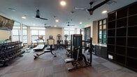 This fitness center offers a thorough and thoughtfully organized workout environment across a single open floor. The space is divided into distinct functional zones, allowing users to transition seamlessly between cardio training, strength work, and functional fitness. The flooring throughout consists of multi-toned carpet tiles, providing a durable and comfortable surface underfoot.

The cardio zone lines the windowed perimeter, where multiple treadmills, ellipticals, and stair climbers are positioned to take advantage of natural light from large windows. Ceiling fans are distributed throughout the space, complementing the recessed lighting to maintain airflow and comfort during workouts.

The strength training area features a full rack of dumbbells, multiple Matrix-brand selectorized cable and weight machines, and a dedicated adjustable bench. A functional training station stores medicine balls, resistance tools, and accessories. Large wall mirrors expand the sense of space and allow users to monitor their form.

A built-in dark-finished shelving unit and a counter with cabinetry provide storage and utility. Wall-mounted flat-screen televisions are positioned throughout, enhancing the workout experience. The overall design is modern and clean, creating a professional, gym-quality atmosphere.