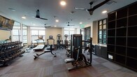 This fitness center offers a thorough and thoughtfully organized workout environment across a single open floor. The space is divided into distinct functional zones, allowing users to transition seamlessly between cardio training, strength work, and functional fitness. The flooring throughout consists of multi-toned carpet tiles, providing a durable and comfortable surface underfoot.

The cardio zone lines the windowed perimeter, where multiple treadmills, ellipticals, and stair climbers are positioned to take advantage of natural light from large windows. Ceiling fans are distributed throughout the space, complementing the recessed lighting to maintain airflow and comfort during workouts.

The strength training area features a full rack of dumbbells, multiple Matrix-brand selectorized cable and weight machines, and a dedicated adjustable bench. A functional training station stores medicine balls, resistance tools, and accessories. Large wall mirrors expand the sense of space and allow users to monitor their form.

A built-in dark-finished shelving unit and a counter with cabinetry provide storage and utility. Wall-mounted flat-screen televisions are positioned throughout, enhancing the workout experience. The overall design is modern and clean, creating a professional, gym-quality atmosphere.