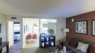 This single-floor unit presents an open and functional layout that connects the living and dining areas in a cohesive, well-organized manner. The living area features a rich chocolate-brown accent wall that serves as a striking focal point, adding warmth and visual contrast against the neutral tones of the surrounding walls and textured ceiling. A plush area rug anchors the seating zone, where a dark sofa and glass-topped coffee table contribute to the space's contemporary aesthetic. A stylish open-cube shelving unit displays decorative vases, framed photos, and sculptural accents, adding character and personalization to the room. A classic floor lamp provides ambient lighting, complementing the overall warmth of the space. Through an open passageway, the dining area is visible and features light hardwood-style flooring, a dining table set with bold red-orange chairs that introduce a vibrant pop of color, and wall art that reinforces the modern design theme. The kitchen, partially visible beyond the dining area, benefits from natural light through a window with a striped valance. Together, the spaces offer a cohesive, modern living environment with thoughtful design details throughout.