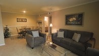 This single-floor residence offers a cohesive and comfortable layout with a warm, inviting ambiance throughout. The living room serves as the central gathering space, furnished with a plush dark gray sofa and matching armchair, each accented with light-colored throw pillows. A glass-top coffee table anchors the seating arrangement, adding a touch of elegance. Crown molding lines the ceiling, lending a classic architectural detail to the room, while recessed lighting and a ceiling fixture provide ambient illumination. A framed botanical artwork adorns one wall, contributing to the space's refined character.

Adjacent to the living area, a dining space is visible, featuring a glass-top dining table surrounded by metal-framed chairs — a design choice that maintains visual openness between the two areas. A decorative metal étagère adds a stylish storage element nearby. Beyond the dining area, a glimpse of the kitchen reveals warm wood cabinetry and under-cabinet lighting. Neutral-toned carpet in the living area and consistent wall colors throughout create a unified, harmonious feel across the entire single-level floor plan.