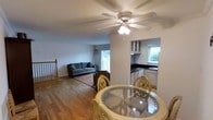 This single-floor space offers an open and functional layout that seamlessly connects the living, dining, and kitchen areas. The living room is a comfortable and well-lit space, featuring a ceiling fan with integrated lighting, warm hardwood flooring, and a window with an air conditioning unit that allows for natural light and ventilation. A sofa and area rug anchor the seating area, which opens to what appears to be a balcony or outdoor deck through a sliding glass door.

The dining area is positioned centrally, furnished with a round glass-top table surrounded by ornate wooden chairs, making it an inviting space for meals and gatherings. Hardwood flooring extends throughout, providing a cohesive and warm aesthetic across the main living spaces.

The kitchen, visible through an open passthrough, is equipped with white cabinetry featuring glass-front upper cabinets, dark countertops, and a sink. A window above the sink brings in additional natural light, brightening the workspace. The layout allows for easy interaction between the kitchen and the adjacent living and dining areas, making the overall space well-suited for both everyday living and entertaining.