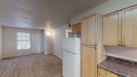This single-floor unit offers a practical and well-organized layout across two bedrooms and one bathroom. The living area is carpeted and receives natural light through a multi-pane window near the front entry door, creating a welcoming and open atmosphere. Textured ceilings are consistent throughout the unit, adding a subtle architectural detail to each room.

The kitchen is efficiently designed with light maple cabinetry extending to the upper walls, providing ample storage space. Dark laminate countertops offer functional workspace, and the kitchen is equipped with a white electric range with overhead ventilation and a full-size refrigerator. A stainless steel sink is positioned beneath a window, allowing natural light into the cooking area. Tile flooring grounds the kitchen space.

Both bedrooms are carpeted and feature neutral walls, windows with blinds for natural light and privacy, and closet space. The layout of the unit, as seen in the overhead view, shows a centrally positioned kitchen that connects fluidly to the living and bedroom areas.

The bathroom includes a light wood vanity with dark countertop, a large mirror with overhead lighting, a tub/shower combination, and tile flooring, offering a clean and functional design consistent with the rest of the unit.