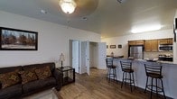 This single-floor space is designed with an open-concept layout that seamlessly connects the living area to the kitchen. The living area features warm hardwood-style flooring that extends throughout the space, complemented by white walls and a ceiling-mounted light fixture that provides ample illumination. A spacious closet with bifold doors offers convenient built-in storage directly off the main living area.

The kitchen is integrated into the open floor plan and includes a raised breakfast bar with seating for two, making it ideal for casual dining or entertaining. Wood-toned upper cabinetry and a stainless steel refrigerator add functional character to the kitchen space. The bar-height counter creates a natural visual division between the kitchen and living areas while maintaining the open feel of the overall layout.

The ceilings are notably elevated, contributing to a sense of openness and airiness throughout the space. The neutral color palette of white walls and warm wood tones creates a cohesive, welcoming ambiance. The efficient single-floor design ensures easy navigation between all areas of the space.