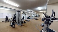This fitness center offers a thorough and well-organized workout environment designed to accommodate a wide range of exercise needs. The space is generously sized, featuring wall-to-wall carpet flooring throughout and a textured ceiling fitted with bright fluorescent lighting that ensures the entire area is evenly illuminated.

The strength training zone is anchored by a multi-station cable machine, complemented by a dedicated weight rack with dumbbells and kettlebells, a flat bench, and step platforms for aerobic exercises. A heavy punching bag suspended from the ceiling adds a dynamic element to the training options available.

The cardio section is well-stocked with multiple treadmills, a stationary upright bike, an elliptical trainer, and a True fitness recumbent machine, providing ample options for cardiovascular workouts. Stability balls are also on hand for core and flexibility training.

Large mirrors are mounted along one wall, visually expanding the space and allowing users to monitor their form during exercise. A wall-mounted flat-screen television is positioned near vertical blind-covered windows and a glass door, offering natural light and views to the exterior. A wall clock and informational signage complete the functional layout, making this fitness center a comprehensive and practical facility for daily use.