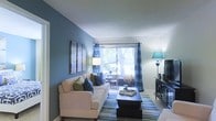 This single-floor residence offers a thoughtfully designed living space that balances comfort and style. The living room serves as the centerpiece of the home, featuring a striking deep blue accent wall that adds depth and personality to the space. A large sliding glass door floods the room with natural light and offers a pleasant view of lush greenery outside, while coordinating blue plaid curtains frame the opening with a polished, cohesive look. The living area is furnished with a comfortable white sofa accented by navy and green throw pillows, complemented by a dark wood coffee table and a striped area rug in coordinating blue and green tones. A flat-screen television and media console anchor one side of the room, while wall art and warm ambient lighting from coordinating lamps enhance the inviting atmosphere. Adjacent to the main seating area, a dining space is visible through the glass door, offering a seamless indoor-outdoor connection. The soft, neutral ceiling and light-toned flooring provide a clean contrast to the bold color palette, contributing to an overall ambiance that feels both vibrant and comfortable. This space is ideal for those seeking a stylish, well-lit living environment with a cohesive design aesthetic.