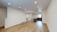 This two-floor space showcases a clean, modern aesthetic defined by an open-concept main level that seamlessly integrates the living area and kitchen. Light-toned hardwood flooring runs throughout, contributing warmth and continuity to the interior. The ceiling features recessed lighting arranged to evenly illuminate the expansive floor plan, with a subtly curved architectural detail adding a distinctive design element overhead.

The kitchen is equipped with dark-toned cabinetry, a central island with a contrasting countertop, and black appliances, including a refrigerator. A window above the sink area allows natural light to filter into the kitchen zone. The cabinetry offers ample storage, and the layout supports an efficient workflow.

A dedicated laundry area is visible within the space, housing a washing machine and providing practical in-unit utility. Several white panel doors lead to additional rooms or storage areas, suggesting versatile use of the floor plan. The crisp white walls throughout enhance the sense of openness and brightness. Overall, the space presents a functional and contemporary environment suited for a variety of uses.