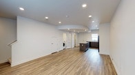 This two-floor space showcases a clean, modern aesthetic defined by an open-concept main level that seamlessly integrates the living area and kitchen. Light-toned hardwood flooring runs throughout, contributing warmth and continuity to the interior. The ceiling features recessed lighting arranged to evenly illuminate the expansive floor plan, with a subtly curved architectural detail adding a distinctive design element overhead.

The kitchen is equipped with dark-toned cabinetry, a central island with a contrasting countertop, and black appliances, including a refrigerator. A window above the sink area allows natural light to filter into the kitchen zone. The cabinetry offers ample storage, and the layout supports an efficient workflow.

A dedicated laundry area is visible within the space, housing a washing machine and providing practical in-unit utility. Several white panel doors lead to additional rooms or storage areas, suggesting versatile use of the floor plan. The crisp white walls throughout enhance the sense of openness and brightness. Overall, the space presents a functional and contemporary environment suited for a variety of uses.