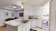 This single-story residence offers a well-designed kitchen that serves as the heart of the home. The kitchen is outfitted with crisp white cabinetry that extends to the upper walls, providing generous storage throughout. Light-toned countertops run along the perimeter and continue onto a peninsula that features a double stainless steel sink, creating a natural divide between the kitchen and the adjoining dining area. This peninsula also functions as a casual gathering or prep space. Appliances include a white dishwasher and a white range, maintaining a cohesive, clean aesthetic throughout the space. Light tile flooring adds a bright, easy-to-maintain surface underfoot, complemented by a decorative navy and white accent rug. A pendant light hangs above the peninsula, adding focused task lighting. The kitchen opens directly into the dining area, which is partially visible through the passthrough layout, featuring a tray ceiling detail, warm wall art, and a dining set with decorative blue glassware. The overall ambiance is clean, functional, and inviting.