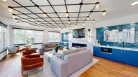 Located at 1031 N Vermont St in Arlington, VA, this thoughtfully designed lounge space makes a strong architectural statement from the moment you enter. The single-floor layout is generously proportioned, offering an open and airy atmosphere ideal for gathering and socializing.

The centerpiece of the main lounge area is a dramatic geometric grid ceiling fitted with an array of pendant lights, casting a warm, even glow throughout the space. Anchoring one wall is a sleek linear fireplace framed by a floor-to-ceiling marble surround, flanked by bold cobalt blue cabinetry with built-in appliances — a vibrant design choice that adds energy and personality to the room. A large wall-mounted television completes this feature wall.

Seating areas are arranged thoughtfully around a white coffee table, with a mix of grey sofas, rust-orange accent chairs, and patterned throw pillows that blend comfort with contemporary style. Warm-toned hardwood flooring runs throughout, adding natural texture and cohesion.

Large windows line the perimeter, flooding the interior with natural light and offering open outdoor views. A secondary alcove area features a striking wood-paneled wall with a textured tile accent installation and a built-in bench, providing a quieter retreat. Curated artwork — including transit and architectural photography — adds a local, urban character to the overall design.
