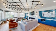 Located at 1031 N Vermont St in Arlington, VA, this thoughtfully designed lounge space makes a strong architectural statement from the moment you enter. The single-floor layout is generously proportioned, offering an open and airy atmosphere ideal for gathering and socializing.

The centerpiece of the main lounge area is a dramatic geometric grid ceiling fitted with an array of pendant lights, casting a warm, even glow throughout the space. Anchoring one wall is a sleek linear fireplace framed by a floor-to-ceiling marble surround, flanked by bold cobalt blue cabinetry with built-in appliances — a vibrant design choice that adds energy and personality to the room. A large wall-mounted television completes this feature wall.

Seating areas are arranged thoughtfully around a white coffee table, with a mix of grey sofas, rust-orange accent chairs, and patterned throw pillows that blend comfort with contemporary style. Warm-toned hardwood flooring runs throughout, adding natural texture and cohesion.

Large windows line the perimeter, flooding the interior with natural light and offering open outdoor views. A secondary alcove area features a striking wood-paneled wall with a textured tile accent installation and a built-in bench, providing a quieter retreat. Curated artwork — including transit and architectural photography — adds a local, urban character to the overall design.