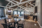 Located at 35101 Harper Ave in Clinton Township, Michigan, this single-floor community clubhouse offers a well-appointed gathering space designed for comfort and social interaction. The architectural centerpiece is the dramatic vaulted ceiling adorned with bold white geometric truss work set against a dark backdrop, lending a modern yet inviting character to the main lounge area.

The expansive main lounge features a painted brick fireplace flanked by large windows that frame wooded outdoor views, filling the space with natural light. A wall-mounted TV above the fireplace and plush seating arrangements centered on a circular area rug create an ideal setting for relaxation and gatherings. Pendant lighting and recessed track lighting complement the overall ambiance.

Adjacent to the lounge, a dedicated coffee bar with a sleek dark countertop and bar stools provides a casual spot for residents to gather. The space transitions smoothly into additional seating areas with bistro-style tables and chairs, as well as a game room equipped with a pool table and pendant lighting, all finished with light hardwood-style flooring.

Throughout the clubhouse, neutral gray walls, patterned carpet tiles, and thoughtful furnishings contribute to a cohesive, contemporary aesthetic. The layout encourages both lively social activity and quiet relaxation in an accessible, single-level design.