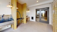 Located at 3903 Cedar Grove Pkwy in Eagan, MN, this purpose-built pet grooming and wash room is a well-appointed, compact amenity space designed with both function and style in mind. The room's most striking visual element is the bold column of golden-yellow ceramic subway tiles, which anchors the grooming station and adds a vibrant, modern contrast to the otherwise neutral white walls and light carpeted flooring. A stainless steel pet wash tub with an accessible ramp provides a dedicated bathing area, accompanied by a professional-grade hose and blow dryer mounted directly to the tiled wall, illuminated by an industrial-style yellow pendant lamp. Adjacent to the grooming station, a built-in light wood cabinetry unit houses a utility sink with a modern faucet, offering a convenient handwashing and prep area. A wall-mounted pet treat or supply dispenser is also integrated into this nook. A large picture window floods the space with natural light and offers views of the surrounding greenery, creating a calm environment. Glass double doors provide easy access. A separate storage area features a full bank of yellow ventilated metal lockers set against charming dog-patterned wallpaper, providing ample personal storage for pet supplies and accessories.