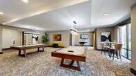 Located in Apopka, FL, this well-appointed game room delivers an inviting recreational experience across a generously sized single-floor layout. The space centers around two full-size pool tables with rich walnut-toned wooden bases, complemented by a foosball table and a wall-mounted flat-screen television for added entertainment. Crystal linear pendant chandeliers hang from a tray ceiling, casting a warm, polished glow across the patterned carpet that anchors the room with an abstract, earthy design.

The neutral gray walls provide a refined backdrop for a curated selection of framed artwork, adding a contemporary artistic flair throughout. A dedicated seating nook features bold yellow accent chairs paired with a compact table, creating an intimate social corner ideal for spectators or casual conversation. Additional high-top bar seating with bar-height chairs is available along one wall, further enhancing the social atmosphere. A cue rack and Connect Four game are also present for varied entertainment options.

Large floor-to-ceiling windows and sliding glass doors line portions of the perimeter, flooding the interior with natural light and offering views of the surrounding greenery. Lush tropical potted plants are strategically placed to introduce vibrant natural elements. The overall ambiance is modern and social, blending upscale design details with a functional, activity-driven layout.