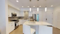 This well-appointed kitchen showcases a clean, contemporary design characterized by crisp white shaker-style cabinetry that extends to the ceiling, providing generous storage throughout. A mosaic tile backsplash in neutral tones of gray and beige adds texture and visual interest behind the professional-grade stainless steel range, complementing the overall color palette. The kitchen is equipped with high-end stainless steel appliances, including a large French door refrigerator and a multi-burner gas range with an integrated oven. A central island with a smooth white countertop serves as both a prep area and a casual dining space, accommodating seating along one side. Elegant cylindrical pendant lights hang above the island, working in tandem with recessed ceiling lighting to create a bright, well-lit environment. Light hardwood flooring runs throughout, adding warmth to the otherwise cool tones of the space. A stainless steel sink is positioned beneath upper cabinetry along the perimeter, completing a highly functional and visually cohesive layout ideal for both everyday cooking and entertaining.