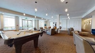 This single-floor amenity lounge is a well-appointed communal space designed for both relaxation and socializing. The main gathering area is generously proportioned, anchored by a full-size pool table and illuminated by a series of elegant crystal pendant lights that add a touch of sophistication to the room. Plush gray lounge chairs and intimate seating arrangements are thoughtfully distributed throughout, offering flexible spaces for casual conversation or quiet relaxation. A dedicated lounge nook features rich teal sofas set against a decorative patterned accent wall, creating a cozy and visually striking retreat within the larger space. Large floor-to-ceiling windows and glass double doors flood the interior with natural light and provide direct access to an outdoor patio, where additional seating is available amid open-air surroundings. The fully equipped kitchen is a standout feature, showcasing dark wood-grain cabinetry, stainless steel appliances including a double wall oven and French door refrigerator, a white quartz countertop, and a spacious bar-height island lined with industrial-style wood-seat barstools. Light tile flooring in the kitchen transitions to warm patterned carpet throughout the lounge areas, tying the space together with a cohesive, contemporary aesthetic.
