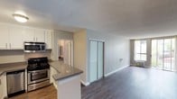 This single-floor space offers an open-concept layout that seamlessly connects the kitchen and living area, creating a functional and cohesive living environment. The kitchen is equipped with stainless steel appliances, including a gas range and built-in microwave, and features white shaker-style cabinetry paired with a gray quartz peninsula countertop that doubles as a casual dining or prep surface. The flooring transitions from lighter tones in the kitchen to rich, dark hardwood-style planks in the living area, adding visual depth and contrast throughout the space.

The living area is bright and open, benefiting from natural light through a window along the far wall. A set of sleek, frosted-glass sliding closet doors provides generous storage while contributing a contemporary aesthetic. Soft gray walls and white baseboards complement the neutral palette, lending the space a clean, modern feel. A textured popcorn ceiling is present throughout. A hallway visible from the main living area leads to additional rooms, suggesting a practical and well-organized floor plan overall.