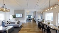 This thoughtfully designed single-floor space offers an open, flowing layout that seamlessly connects multiple functional areas. The living area is anchored by a wall-mounted flat-screen TV and furnished with a contemporary light gray sofa, complemented by decorative throw pillows and a potted fiddle-leaf fig plant that adds a touch of greenery. Warm-toned hardwood flooring runs throughout, unifying the space with a sense of continuity and warmth.

A standout architectural feature is the elegant coffered ceiling, which adds visual depth and a refined character to the main living zone. The dining area is situated adjacent to the living space and features a sleek dining table surrounded by dark upholstered chairs, illuminated by a stylish pendant light fixture overhead.

Beyond the main living and dining areas, a connecting corridor leads to a secondary lounge or sitting room, visible in the background, which is furnished with additional seating and accented by a dark teal feature wall that introduces a bold pop of color. Framed artwork and careful lighting selections throughout contribute to a polished, gallery-like ambiance. Track lighting and recessed fixtures ensure the space is well-lit, while sheer window treatments allow natural light to filter in softly, enhancing the bright and airy atmosphere overall.