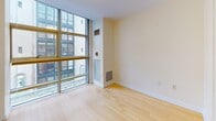 Located at 631 Washington St in Boston, MA, this single-floor unit offers a thoughtfully arranged open layout bathed in natural light. The living area is a standout feature, anchored by expansive floor-to-ceiling windows with a gridded frame that frame views of the surrounding urban streetscape, filling the space with daylight and creating a strong architectural focal point. Light-toned hardwood flooring runs throughout the main living areas, lending warmth and visual continuity.

The kitchen is well-equipped with white cabinetry extending to the ceiling, providing ample storage. White appliances—including a refrigerator, range, microwave, and dishwasher—complement the clean aesthetic. A double sink and neutral countertops complete the functional workspace. The kitchen flows openly into the living area, making the overall layout feel connected and spacious.

The bathroom is finished with cream-toned square wall and floor tiles, featuring a combined soaking tub and shower with chrome fixtures. A white vanity with cabinet storage, integrated sink, and a mirrored medicine cabinet round out the practical amenities. The unit's neutral palette and recessed lighting throughout contribute to a bright, clean, and cohesive overall ambiance.