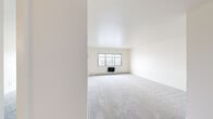 This 1-bedroom, 1-bathroom unit offers a clean and functional layout across a single floor. The interior is finished with neutral white walls and consistent grey carpet throughout the living, bedroom, and hallway areas, creating a cohesive and bright atmosphere. The spacious living room features multi-pane windows that invite generous natural light, complemented by baseboard heating along the perimeter walls. A pass-through opening connects the living area to the kitchen, which is equipped with warm wood-toned cabinetry and hard surface flooring, adding a touch of contrast to the otherwise light palette. The bedroom is a comfortable, well-proportioned space with its own window offering outdoor views, baseboard heating, and a built-in closet with a wood-panel sliding door and hanging rod for ample storage. A hallway with hardwood-style flooring connects the main living areas to the full bathroom, which features classic tile flooring in a small grid pattern, a vanity with a medicine cabinet mirror, and a shower with a curtain enclosure. The layout is practical and efficient, making effective use of the available square footage throughout.