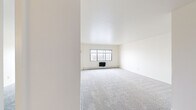 This 1-bedroom, 1-bathroom unit offers a clean and functional layout across a single floor. The interior is finished with neutral white walls and consistent grey carpet throughout the living, bedroom, and hallway areas, creating a cohesive and bright atmosphere. The spacious living room features multi-pane windows that invite generous natural light, complemented by baseboard heating along the perimeter walls. A pass-through opening connects the living area to the kitchen, which is equipped with warm wood-toned cabinetry and hard surface flooring, adding a touch of contrast to the otherwise light palette. The bedroom is a comfortable, well-proportioned space with its own window offering outdoor views, baseboard heating, and a built-in closet with a wood-panel sliding door and hanging rod for ample storage. A hallway with hardwood-style flooring connects the main living areas to the full bathroom, which features classic tile flooring in a small grid pattern, a vanity with a medicine cabinet mirror, and a shower with a curtain enclosure. The layout is practical and efficient, making effective use of the available square footage throughout.