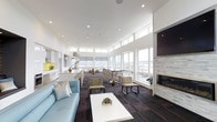 This contemporary single-floor space is designed with an open-concept layout that maximizes both functionality and visual appeal. Soaring ceilings and a continuous band of large clerestory windows along the upper walls flood the interior with abundant natural light, creating an airy and expansive atmosphere throughout.

The space is thoughtfully divided into distinct yet seamlessly connected zones. A fully equipped modern kitchen anchors one end, featuring sleek white cabinetry, stainless steel appliances, and pendant lighting over a generous island. Adjacent to the kitchen, a long bar-height counter with stylish stools serves as an informal dining and gathering area.

The central zone features additional casual seating with armchairs and tables arranged to encourage conversation and collaboration. Toward the opposite end, a comfortable lounge area is anchored by a light blue sofa and a clean-lined white coffee table, offering a relaxed setting for unwinding. A wall-mounted flat-screen television and an elegant tiled fireplace with a hearth serve as focal points in this lounge area, adding warmth and a sophisticated touch.

Dark hardwood-style flooring with a subtle tile pattern grounds the space, providing a rich contrast to the bright white walls and ceilings. Large windows along the exterior walls offer generous views of the surrounding outdoor environment, further enhancing the open, welcoming character of this well-appointed space.