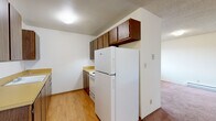 This single-floor residence offers a straightforward and functional layout designed for comfortable everyday living. The galley-style kitchen is a standout feature, lined on both sides with warm wood-toned cabinetry that extends to the upper walls, providing ample storage. Yellow-toned countertops offer generous prep space, complemented by a built-in sink, dishwasher, and a full-size refrigerator. Light hardwood-style flooring runs through the kitchen, transitioning to plush carpeting in the living and sleeping areas. The living room is generously sized, featuring a large window that invites natural light and views of surrounding greenery. A baseboard heating system runs along the walls, ensuring consistent warmth throughout. The entryway includes a closet for convenient storage near the front door. The bedroom is well-proportioned, featuring two windows with blinds, a wall-mounted air conditioning unit, a baseboard heater, and a mirrored sliding-door closet that enhances the sense of space. The full bathroom is cleanly appointed with a tub-shower combination, a wood-vanity with a yellow countertop, tile-style flooring, and a wall-mounted mirror. Textured ceilings are consistent throughout the space, adding a classic residential character.