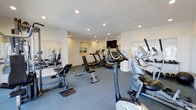 This single-floor fitness center offers a comprehensive workout environment designed to accommodate a range of exercise routines. The space is outfitted with an assortment of cardio equipment, including stationary bikes, treadmills, and elliptical machines, alongside strength training benches and resistance equipment. The layout is thoughtfully organized to allow comfortable movement between machines.

The room features a neutral color palette with white walls and a durable gray flooring surface suited for gym use. Recessed lighting throughout the ceiling provides consistent, even illumination across the entire space. Large mirrors are mounted along one wall, enhancing the sense of openness and allowing users to monitor their form during workouts.

Natural light filters in through windows on one side of the room, contributing to a bright and energizing atmosphere. A wall-mounted flat-screen television is positioned at the far end of the space, offering entertainment during workouts. The overall environment is clean, functional, and well-maintained, making it a practical and inviting facility for daily fitness use.