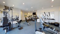 This single-floor fitness center offers a comprehensive workout environment designed to accommodate a range of exercise routines. The space is outfitted with an assortment of cardio equipment, including stationary bikes, treadmills, and elliptical machines, alongside strength training benches and resistance equipment. The layout is thoughtfully organized to allow comfortable movement between machines.

The room features a neutral color palette with white walls and a durable gray flooring surface suited for gym use. Recessed lighting throughout the ceiling provides consistent, even illumination across the entire space. Large mirrors are mounted along one wall, enhancing the sense of openness and allowing users to monitor their form during workouts.

Natural light filters in through windows on one side of the room, contributing to a bright and energizing atmosphere. A wall-mounted flat-screen television is positioned at the far end of the space, offering entertainment during workouts. The overall environment is clean, functional, and well-maintained, making it a practical and inviting facility for daily fitness use.