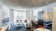 This single-floor urban residence showcases an industrial-modern aesthetic defined by its striking exposed concrete ceiling, which contrasts elegantly with crisp white walls and dark hardwood flooring. The open-plan layout seamlessly integrates the living, dining, and work areas into one cohesive space. Floor-to-ceiling windows flood the interior with natural light while offering expansive city views of the surrounding skyline, creating a strong visual connection to the urban environment outside. The living area is anchored by a light-toned sofa accented with burnt orange and patterned cushions, complemented by a blue area rug and a minimalist twin-table coffee setup. Adjacent to the living space, a compact yet functional workspace features a slim desk, task lighting, and framed wall art. A dining area with vibrant orange chairs and a sculptural gold chandelier adds a bold focal point near the entry. The overall ambiance is sophisticated and contemporary, balancing raw industrial elements with carefully curated furnishings and warm accent tones throughout.