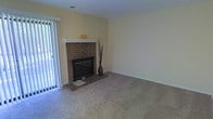 This single-floor residence offers a warm and inviting living environment highlighted by thoughtful details and a comfortable layout. The living room serves as the heart of the space, anchored by a traditional brick fireplace with a clean white mantle that adds classic charm and character. Soft, neutral-toned wall-to-wall carpeting runs underfoot, contributing to the cozy atmosphere of the room. A large sliding glass door fitted with vertical blinds allows generous amounts of natural light to pour in, brightening the pale yellow walls and creating an open, airy feel. The high ceiling further enhances the sense of spaciousness in the room. The overall ambiance is calm and understated, offering a clean, move-in ready canvas with timeless architectural touches. The brick fireplace stands as a clear focal point, lending warmth both visually and functionally to the living area. Together, these elements create a comfortable, welcoming space well-suited for everyday living.