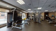 Located at 10 Kings Road in Madison, NJ, this fitness center is a well-appointed, single-floor facility designed to support a full range of exercise routines. The space features a drop-panel ceiling with recessed and flush-mounted lighting that ensures bright, even illumination throughout. Durable rubber flooring covers the entire area, providing traction and impact absorption suited to high-activity use.

The cardio section is equipped with multiple True-brand treadmills and upright stationary bikes, along with elliptical trainers, all positioned near large windows that allow natural light to filter in. A wall-mounted flat-screen TV accompanies the cardio area for entertainment during workouts.

The strength training section includes Cybex dual-cable resistance machines, a multi-functional cable crossover station with TRX suspension straps, and a dedicated free weights area with a tiered dumbbell rack. Adjustable weight benches and additional selectorized machines—including a leg extension/curl unit—round out the strength offerings.

Full-length mirrors line one wall, visually expanding the space and aiding in form monitoring. A textured accent wall near the entry corridor adds a design element to the otherwise clean, neutral aesthetic. The facility also includes a dedicated entry door with keycard access, reinforcing a controlled, professional environment.
