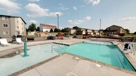 This outdoor community pool area offers a well-maintained aquatic amenity designed for recreational use. The pool itself is generously proportioned, featuring clear, blue-green water and a smooth concrete surround that provides ample deck space for lounging and movement around the perimeter. Entry into the pool is facilitated by stainless steel handrails and steps, making access straightforward and safe for swimmers of varying ages.

The deck area is finished with large concrete pavers, providing a clean and durable surface that extends broadly around the pool. Lounge chairs are positioned along the deck, offering spots to relax in the open air. The entire pool area is enclosed by a black metal fence, providing both security and clear boundary definition. Tall light poles are stationed throughout the space, ensuring the area is well-lit during evening hours.

The setting enjoys open sky exposure, allowing abundant natural sunlight throughout the day. Views from the pool deck extend outward to the surrounding residential neighborhood, including single-family homes and a distinctive barn-style structure with a red roof visible in the background, contributing to a suburban, community-oriented atmosphere.