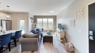 This single-floor residence offers a thoughtfully designed layout with cohesive finishes throughout. The kitchen is a standout feature, showcasing white shaker-style cabinetry extending to the ceiling, a herringbone tile backsplash, and a full suite of stainless steel appliances including a refrigerator, electric range with microwave above, and dishwasher. Light wood-look flooring runs through the kitchen, complemented by pendant lighting and a marble-look countertop island edge for added prep space.

The living room is bright and welcoming, featuring the same light wood flooring, a large window that brings in natural light and outdoor views of surrounding greenery, and access to an exterior balcony. A hallway connects the main living area to the private quarters, maintaining a clean, open flow through the home.

The bedroom is generously sized with plush carpeting, a ceiling fan with light fixture, a closet with louvered doors, and a window dressed with dark curtains. A second bedroom with similar carpeting and ceiling fan provides additional flexibility.

The bathroom is finished with a white vanity, gray quartz-style countertop, a wide framed mirror, and a glass-enclosed walk-in shower with tiled walls, creating a polished, functional space.