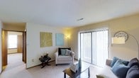 This single-floor residence features a welcoming living room designed with comfort and functionality in mind. The space is finished with wall-to-wall carpeting in a neutral tone, contributing to a warm and inviting atmosphere. A prominent sliding glass door fitted with vertical blinds serves as both a source of natural light and a direct access point to an outdoor balcony, offering views of surrounding greenery. The textured ceiling adds subtle visual interest overhead, while the light-colored walls provide a clean, versatile backdrop. The room accommodates a seating arrangement that includes an upholstered armchair accented with decorative pillows, alongside a dark coffee table and coordinating side table, illustrating the practical use of the available space. Two framed canvas artworks in muted green and blue tones are displayed on the wall, adding a touch of color and personality. A floor lamp positioned near the seating area supplements the natural light entering through the sliding door. The overall design of the living room reflects a straightforward, functional layout with an emphasis on natural light and easy indoor-outdoor connectivity.