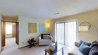 This single-floor residence features a welcoming living room designed with comfort and functionality in mind. The space is finished with wall-to-wall carpeting in a neutral tone, contributing to a warm and inviting atmosphere. A prominent sliding glass door fitted with vertical blinds serves as both a source of natural light and a direct access point to an outdoor balcony, offering views of surrounding greenery. The textured ceiling adds subtle visual interest overhead, while the light-colored walls provide a clean, versatile backdrop. The room accommodates a seating arrangement that includes an upholstered armchair accented with decorative pillows, alongside a dark coffee table and coordinating side table, illustrating the practical use of the available space. Two framed canvas artworks in muted green and blue tones are displayed on the wall, adding a touch of color and personality. A floor lamp positioned near the seating area supplements the natural light entering through the sliding door. The overall design of the living room reflects a straightforward, functional layout with an emphasis on natural light and easy indoor-outdoor connectivity.