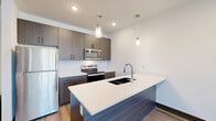 This thoughtfully designed one-bedroom, one-bathroom residence at 8320 N Green Hills Rd in Kansas City, MO offers a clean, contemporary aesthetic across a single floor. The open-concept kitchen and living area serves as the heart of the unit, showcasing dark wood-grain cabinetry, a bold kitchen island with a quartz countertop and undermount sink, and stainless steel appliances including a refrigerator, range, microwave, and dishwasher. Stylish pendant lighting hangs above the island, complementing the recessed lighting and light wood-tone flooring that flows throughout the main living space. The spacious bedroom features plush carpeting, a ceiling fan, and a large window that invites natural light and outdoor views. It connects directly to the full bathroom, which offers a wide vanity with quartz countertops, a large mirror, and wood-tone cabinetry. Two closets provide ample storage, while a dedicated laundry area and utility room add everyday convenience. Throughout the unit, dark door and baseboard trim creates a polished contrast against the bright white walls, lending the space a modern, cohesive character.